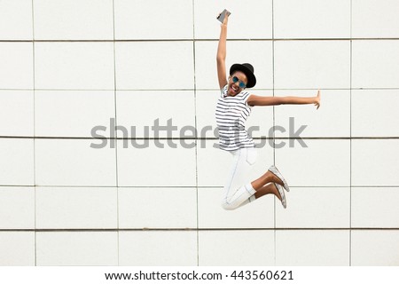 happy modern young black woman with sunglasses, hat and striped shirt  listening music outside and jumping