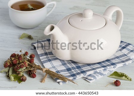  Teapot on a napkin, a cup and sprigs of the dried of wild strawberry on a light wooden background.