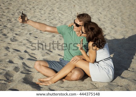 Young loving couple sitting on the sand at the beach and make selfies.