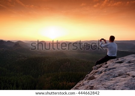 Tourist in grey t-shirt takes photos with smart phone on peak of rock. Dreamy hilly landscape below, spring orange pink misty sunrise in beautiful valley below rocky mountains. Hiking man taking photo