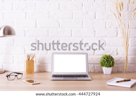 Front view of creative hipster desktop with blank white laptop, smart phone, glasses, lamp, stationery and decorative items on white brick wall background. Mock up