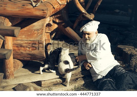 bearded man cook chef in uniform and hat with long beard on smiling face holding iron old tea kettle near black and white cats sunny day outdoor on wooden background