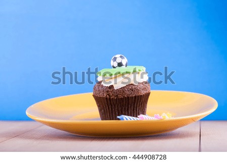 Delicious cupcake with ball icon on it with yellow plate and three candles on wooden desk and blue background