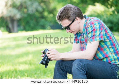 Young handsome guy in colorful shirt looking  on pictures in camera and sitting on stump in park