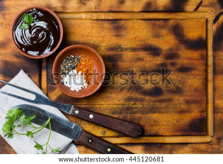Barbecue sauce, salt, red and black pepper in clay bowls, meat fork, knife and napkin on wooden cutting board. Top view. Copy space