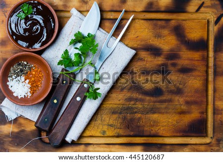 Barbecue sauce, salt, red and black pepper in clay bowls, meat fork, knife and napkin on wooden cutting board. Top view. Copy space