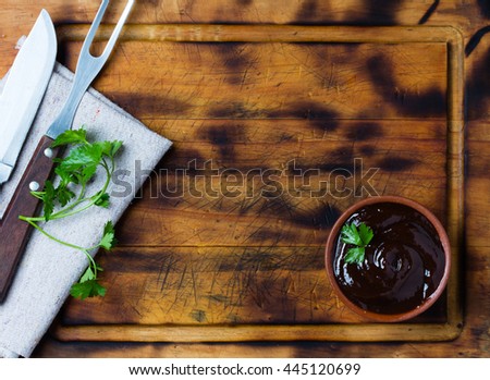 Barbecue sauce in clay bowl, meat fork and knife and napkin on wooden cutting board.
