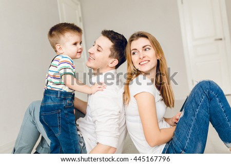 Close-up of family picture of two young parents playing with their boy child. They sit on the floor and father holds his son. They wear white t-shirt and jeans. 