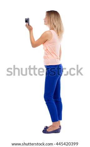 back view of standing young beautiful  woman  and using a mobile phone. girl  watching. Rear view people collection.  backside view person. Blonde in blue pants with one hand removes a compact camera