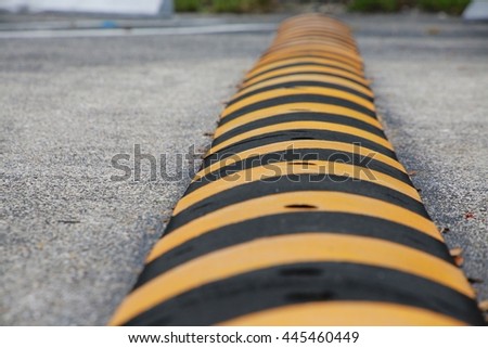 Yellow and Black Striped Speed Bump with Nails Visible Frame Right in Parking Lot with Diagonal Spaces, White Blocks and Green Grass Above in Background