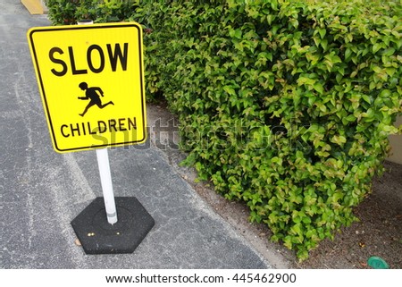 Yellow Slow Children Sign with Black Lettering and Child Crossing Illustration on Small Post on Pavement in Parking Lot Frame Left with Bushes Frame Right