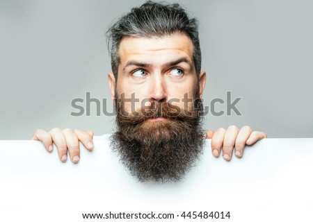 handsome bearded man with long lush beard and moustache on surprised face with white paper sheet in studio on grey background, copy space Royalty-Free Stock Photo #445484014