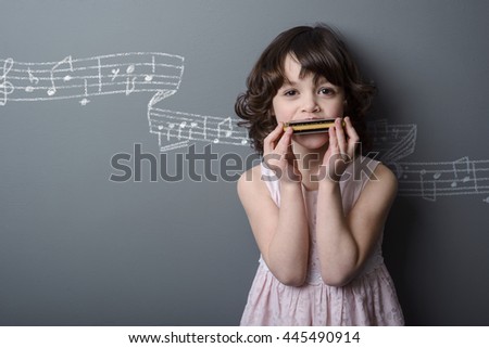 Interesting stave with notes on the neutral background in the studio. Picture painted with a chalk. Amazing little girl plays the harmonica and looks in the camera.