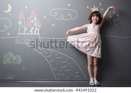 Little princess near her castle smiles and holds her dress with hand. Depicted road, crown and stars on the neutral background. True emotion of happiness of the child. Royalty-Free Stock Photo #445491022