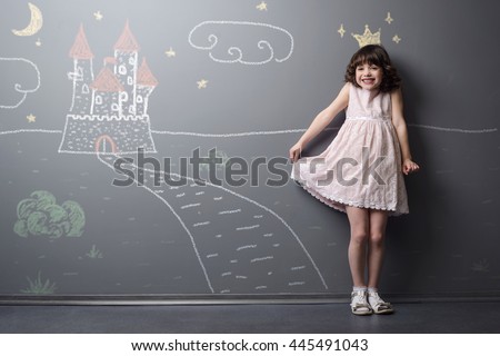 Shy smiling little princess is very pleased and happy to imagine herself the future queen. Depicted with chalk drawing with crown, road and castle on the grey wall.
