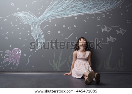 Lovely girl has an adventure to the world of sea. Big blue whale, violet octopus and fish depicted with chalk on the grey wall. Child sits on the floor and looks upwards.