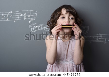 Little musician with cute curls dressed up in pink dress. Pretty girl blows the air in harmonica to produce cool melody. Painted with chalk stave with the notes on the grey wall in the background.