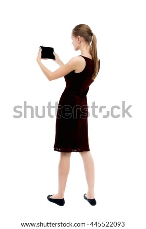 back view of standing young beautiful  girl with tablet computer in the hands of. girl  watching. Rear view people collection.   The blonde burgundy dress holding to a horizontal smartphone.