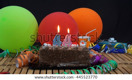 Chocolate birthday cake with candles burning on rustic wooden table with background of colorful balloons, gifts, plastic cups and streamers with black background