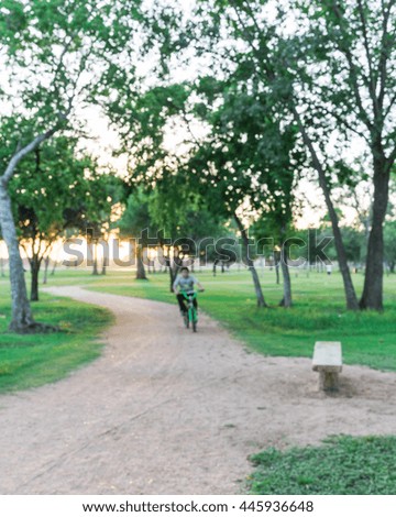Defocused, blurred motion background of a boy riding bike though trail in urban park with bench illuminated by sunshine at sunset in Houston, Texas, US. Kid summer activities background and concept.