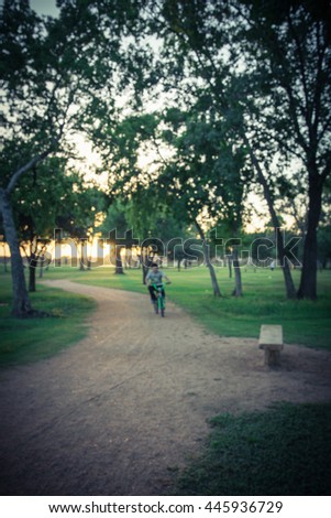 Defocused, blurred motion background of a boy riding bike though trail in urban park with bench illuminated by sunshine at sunset in Houston, Texas, US. Kid summer activities background and concept.