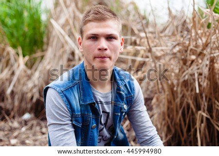 guy in a gray T-shirt and denim jacket sitting in the reeding on the shore of the lake and biting his lip