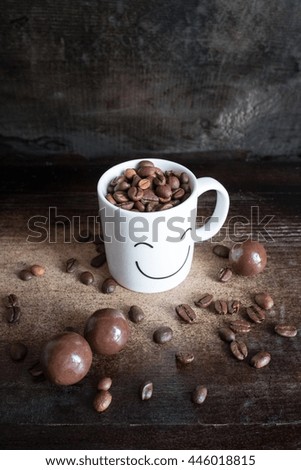 White coffee mug with a smiling picture face, and coffee beans, chocolate candies on dark wooden background