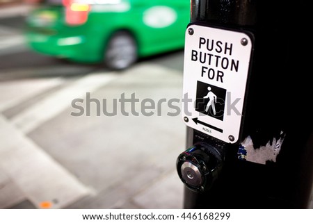 Crosswalk sign and button on a black pole, with green taxi passing in the background