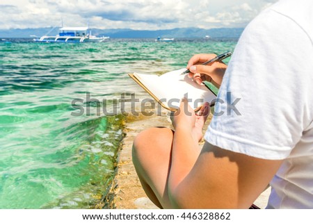 caucasian man is sitting on the breakwater of tropical turquoise sea and writing by pen in note pad with philippine boat background at sunny summer day