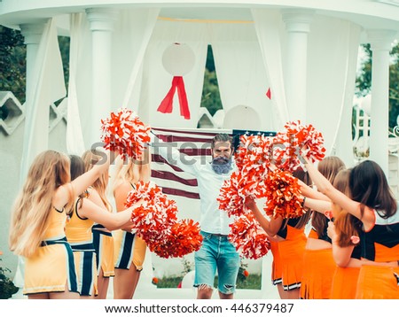 young patriotic happy bearded man with long blue beard holding american flag outdoor celebrating independence day usa with cheerleader team dancing with pompons