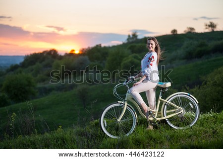 Girl in Ukrainian embroidery with vintage bike looking to the camera. Blurred background with beautiful sunset