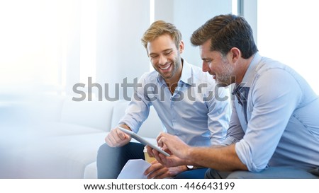 Smiling young businessman discussing something positive with his mature colleague, and using a digital tablet together Royalty-Free Stock Photo #446643199