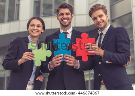 Handsome young businessmen and lady in classic suits are holding colorful pieces of puzzle, looking at camera and smiling, standing outside the office building