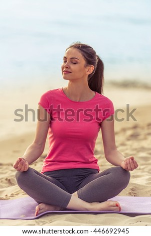 Beautiful girl in sport clothes is meditating and smiling while sitting in lotus position on yoga mat on the beach