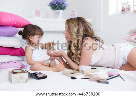 Mother and daughter in white having fun doing lip make-up. Mother and daughter doing makeup sitting on the bed in the bedroom