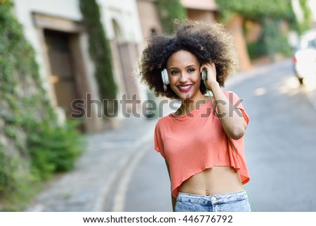 Portrait of young attractive black girl in urban background listening to the music with headphones. Woman wearing leather jacket and blue jeans with afro hairstyle
