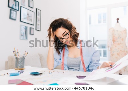 A pretty brunette girl is sitting at the table in the workshop studio. She wears blue shirt and white watch