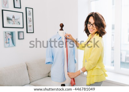 Brunette girl in a yellow jacket  makes  fitting shirt on mannequin. She works in a large workshop studio. She is smiling to the camera.