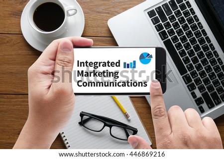 integrated marketing communications    (IMC) message on hand holding to touch a phone, top view, table computer coffee and book