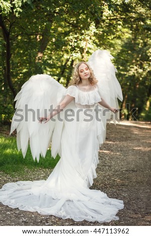 Beautiful blonde girl in a long white dress wears huge white angel wings and walking in the park