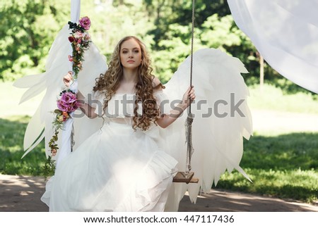 Beautiful blonde girl in a long white dress wears huge white angel wings sitting and daydreaming on a swing