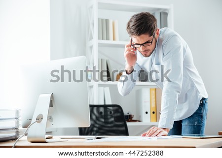 Young handsoome business man standing at desk working on documents with mobile phoone in office