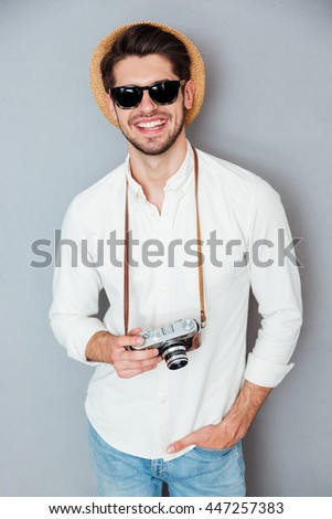 Smiling young man in hat and sunglasses with old vintage camera over grey background
