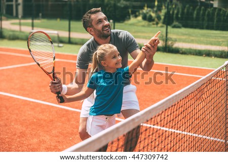 Tennis is fun when father is near. Cheerful father in sports clothing teaching his daughter to play tennis while both standing on tennis court  Royalty-Free Stock Photo #447309742