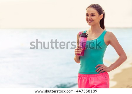 Beautiful girl in sport clothes is holding a bottle of water, looking away and smiling, standing on the beach after workout