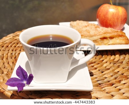 Coffee in a white cup with a purple flower on the side. Apple French pastry turnover and apple in the background