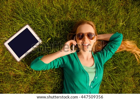 Smiling girl lying on lawn with tablet and talking on phone