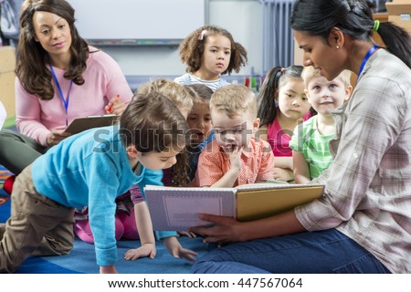Female teacher giving a lesson to nursery students. They are sitting on the floor and there is a teacher taking notes.