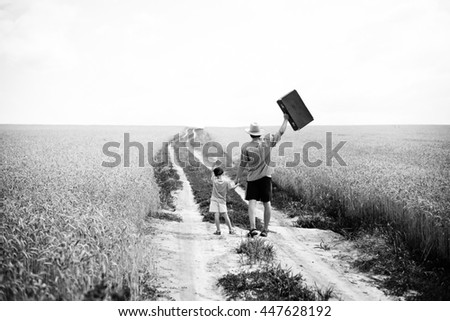 Black and white photography back view of man with child traveling on the countryside rural road. Father in straw hat enjoying walking with his son and raising suitcase up.