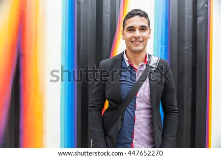 Modern young man with Arab features with shoulder bag and jacket without a tie on multicolored background and the hands in his pocket